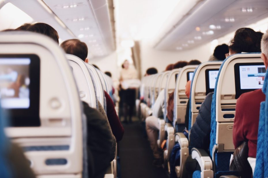 Rows of airline passengers seated in a plane cabin with seat-back screens and a crew member in the aisle.